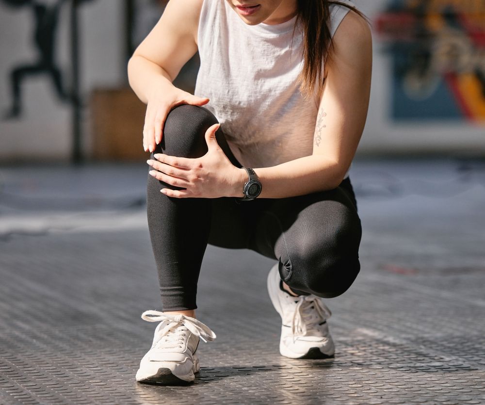 Person kneeling on a gym floor, holding a bent knee in white sneakers and black leggings.