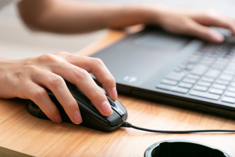 Hands using a black mouse beside a laptop on a wooden desk