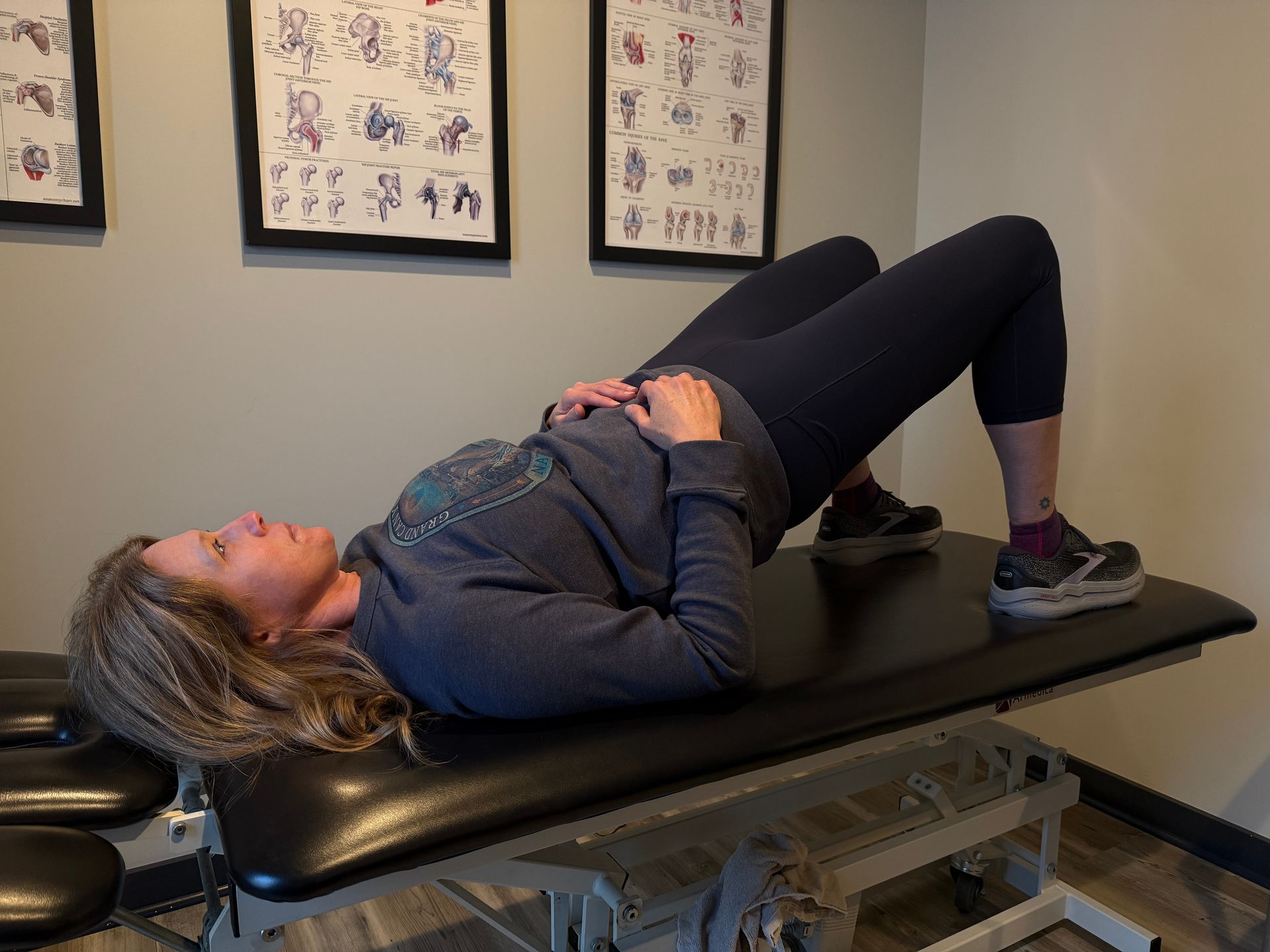 Woman on a table doing a bridge exercise with hands on stomach, in a room with anatomical charts.