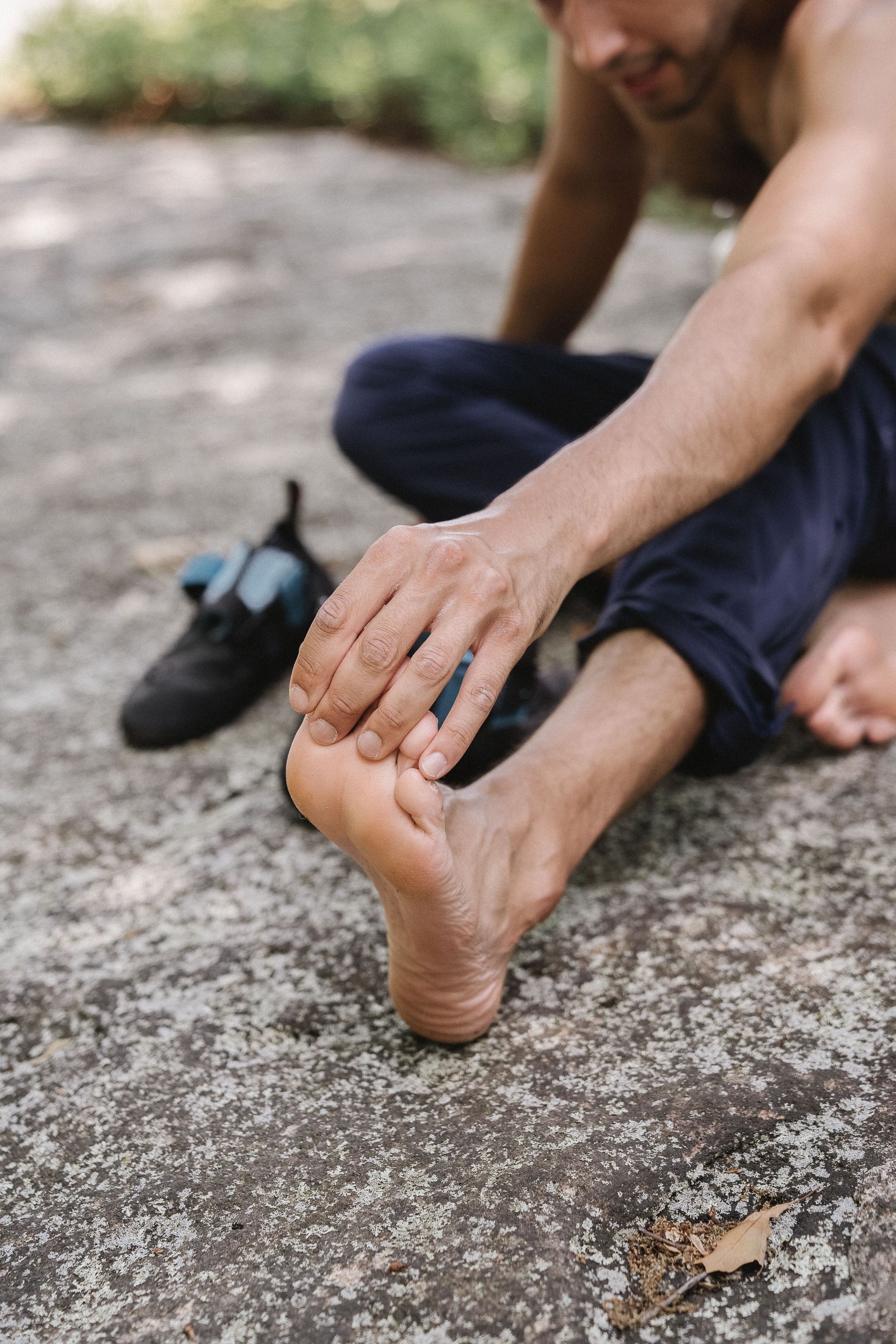 Person sitting outdoors, holding and massaging a bare foot on a rocky ground