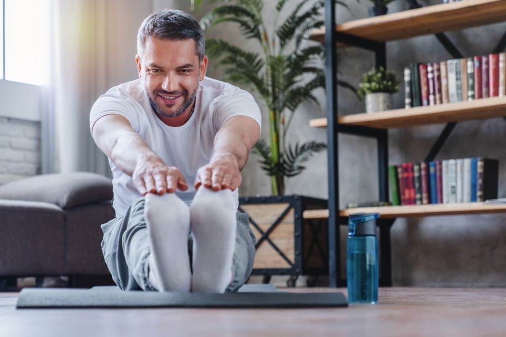 Man stretches, touching toes while sitting on a mat indoors. He smiles, with bookshelf and water bottle nearby.
