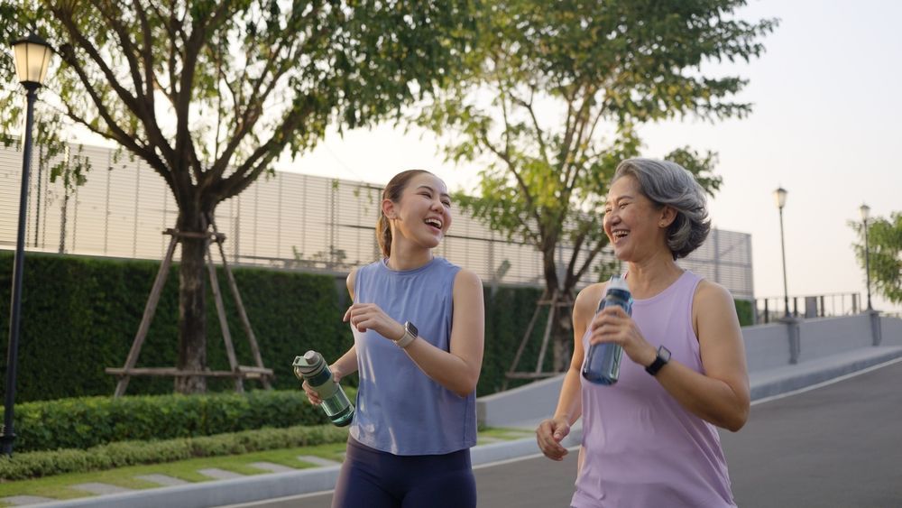 Woman and older woman jogging on a path, laughing and holding water bottles in an outdoor setting.