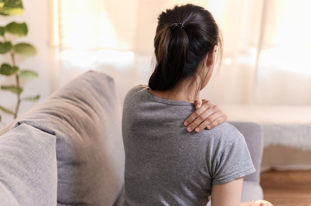Person sitting on a couch, holding their shoulder in a bright living room.