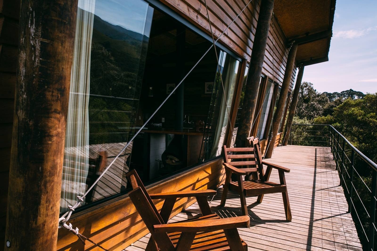 Cadeiras de madeira em uma varanda ensolarada com piso trançado, ao lado de grandes janelas abertas e vista para a vegetação.