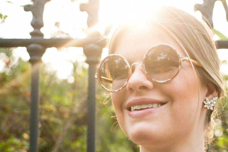 Mulher sorridente com óculos de sol redondos ao ar livre, sob a luz solar intensa e com vegetação ao fundo.