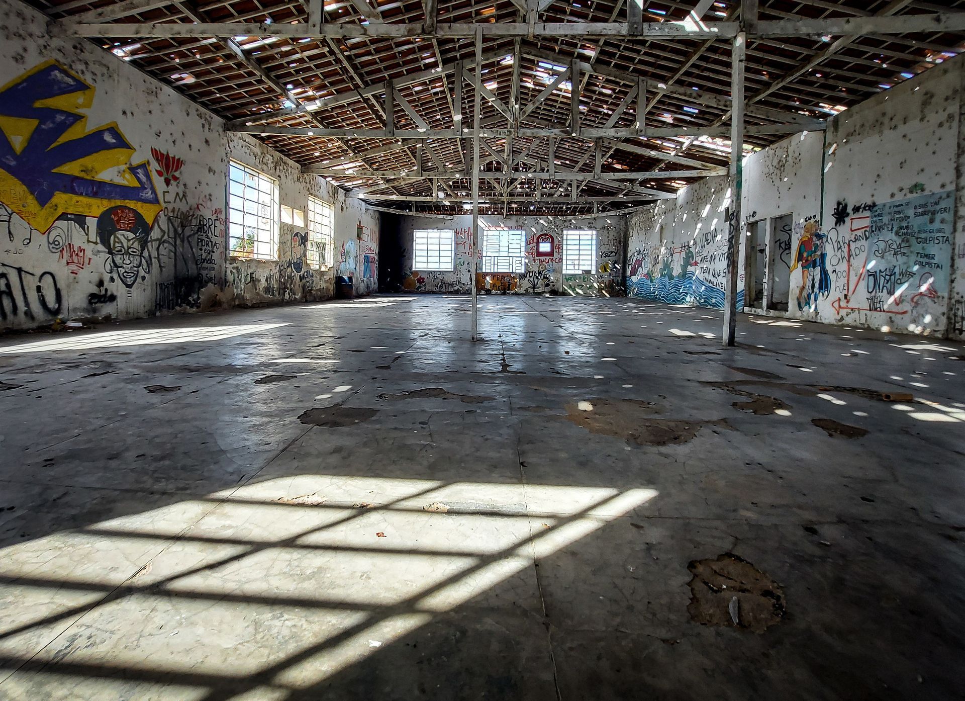Interior de um armazém abandonado, coberto de grafites, com piso de concreto e luz solar entrando pelas aberturas do teto.