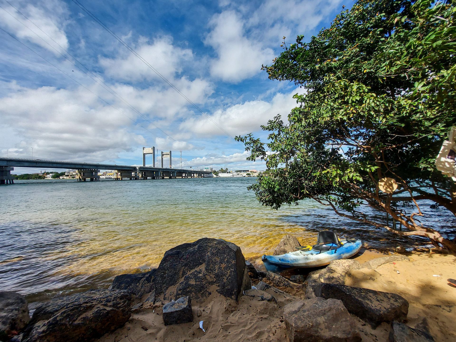 Margem arenosa com caiaque sob uma árvore frondosa, água e ponte sob um céu azul e nublado.