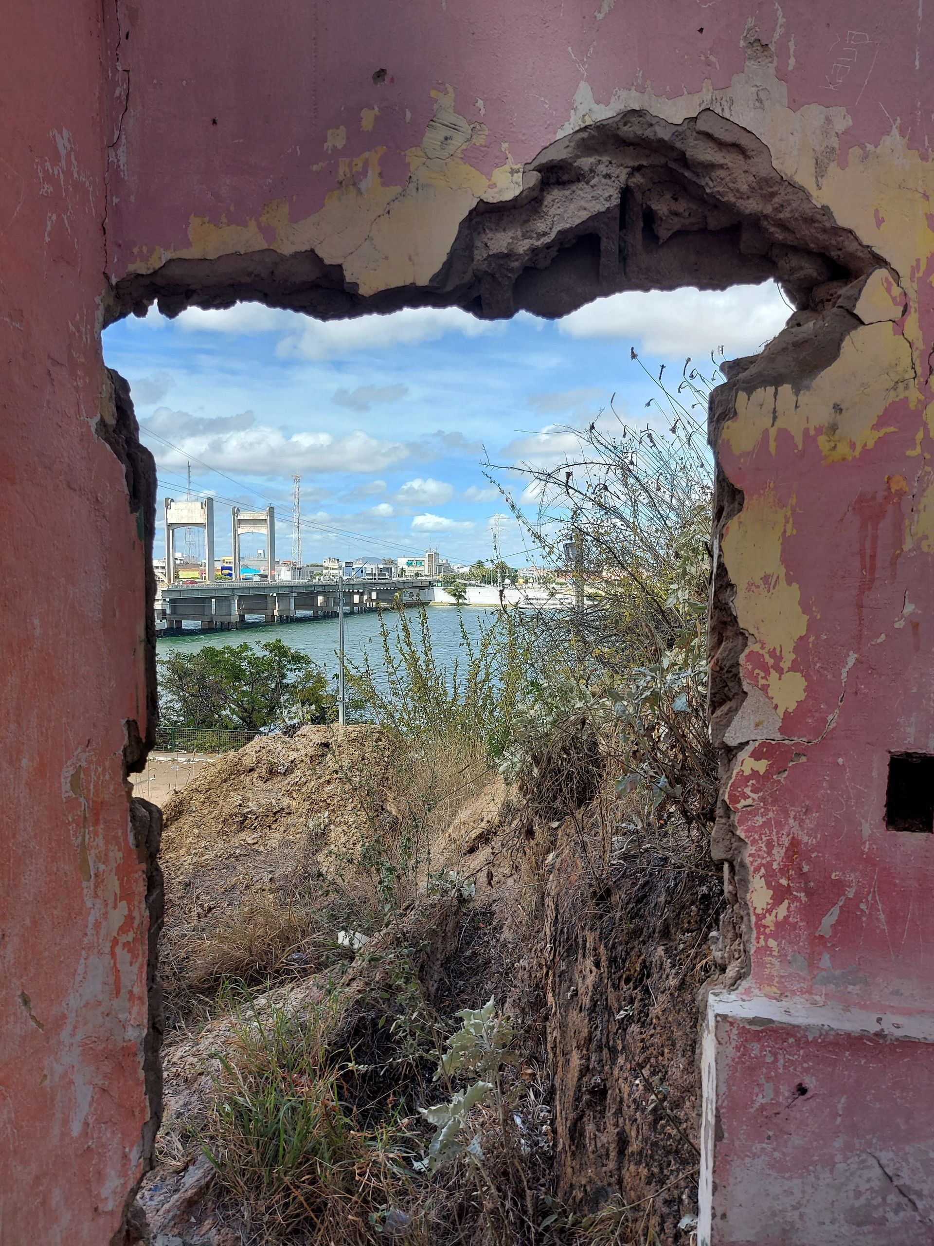 Vista através de uma parede rosa quebrada para um rio, uma ponte e o céu azul além da vegetação rasteira.