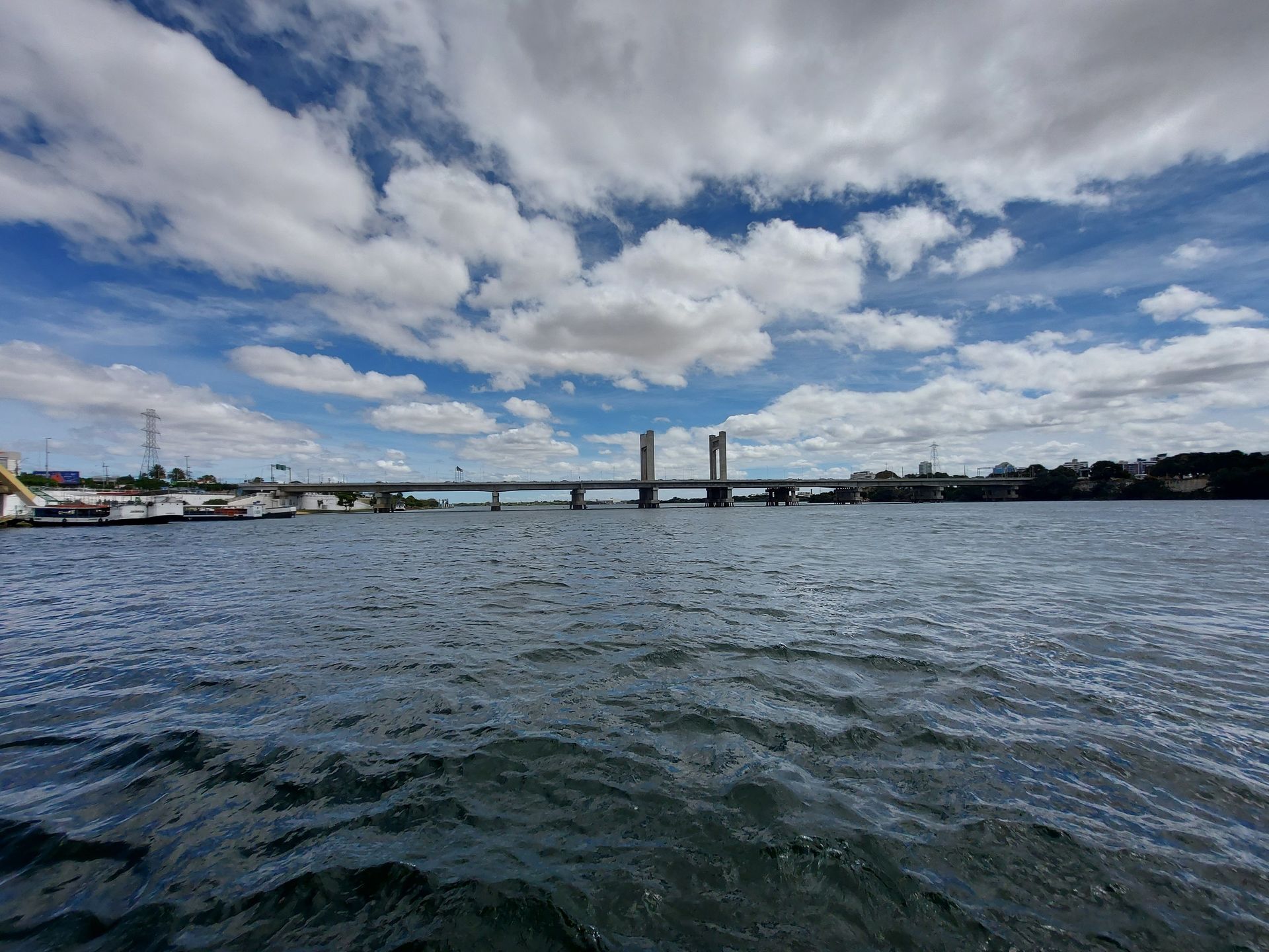 Rio largo com águas agitadas sob um céu nublado, visto em direção a uma ponte com torres gêmeas ao longe.