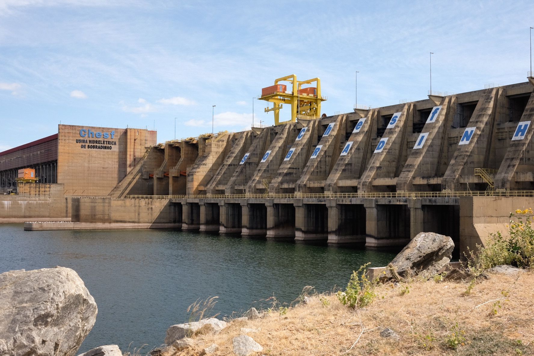 Barragem de concreto com comportas de vertedouro ao lado de um reservatório tranquilo sob um céu azul claro.