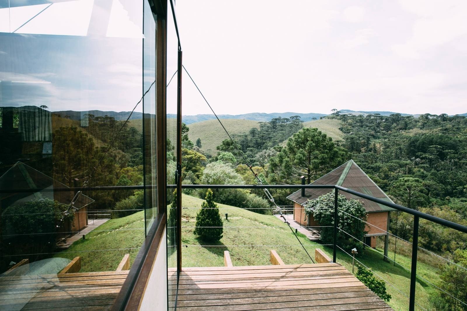 Vista da varanda para colinas verdes e uma casa com telhado de telhas, com guarda-corpo de vidro e deck de madeira.