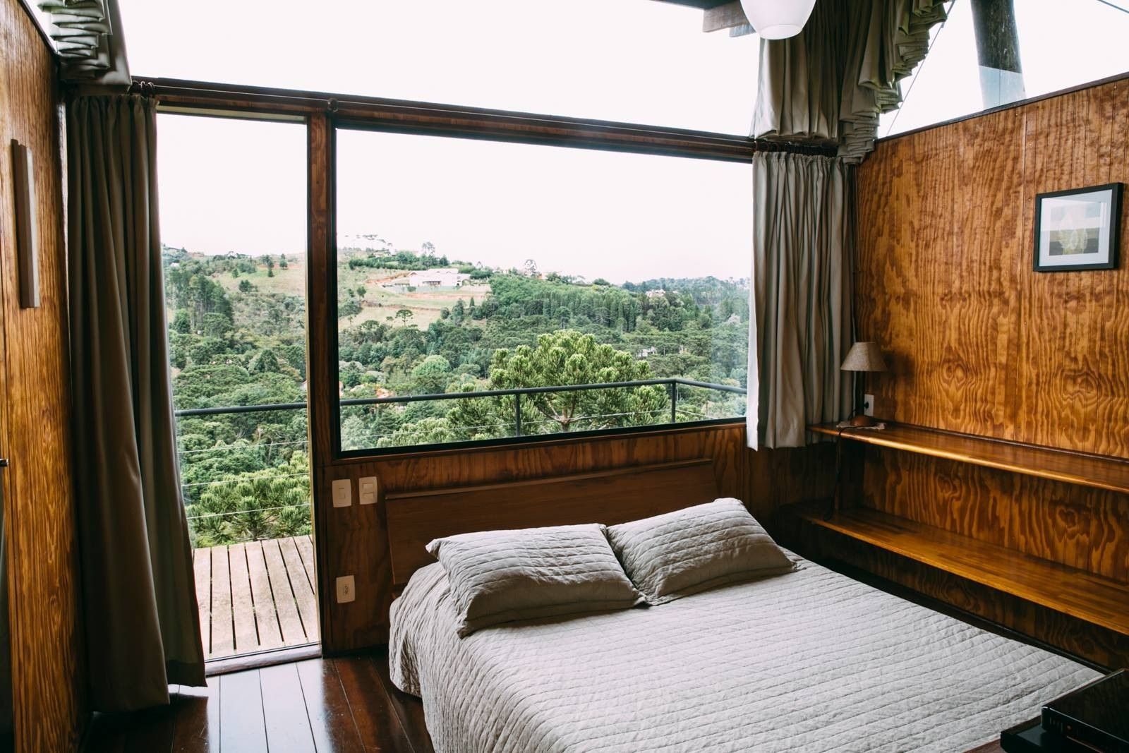 Quarto aconchegante com paredes de madeira e cama de frente para uma grande janela com vista para colinas verdejantes.