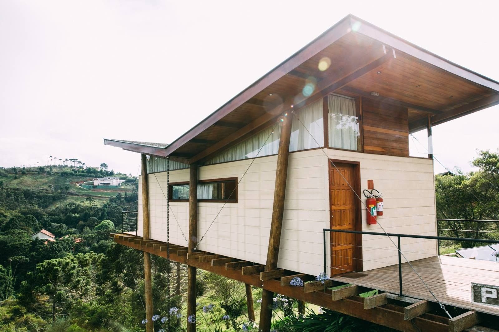 Casa moderna sobre palafitas, com paredes de madeira e brancas, situada numa encosta com vista para um vale verdejante.