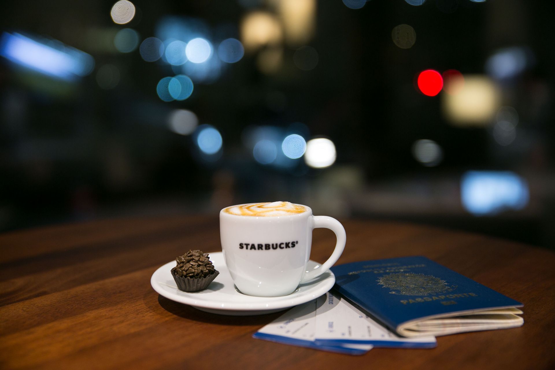 Xícara de café e doces sobre uma mesa ao lado de um caderno azul, com luzes da cidade desfocadas ao fundo.