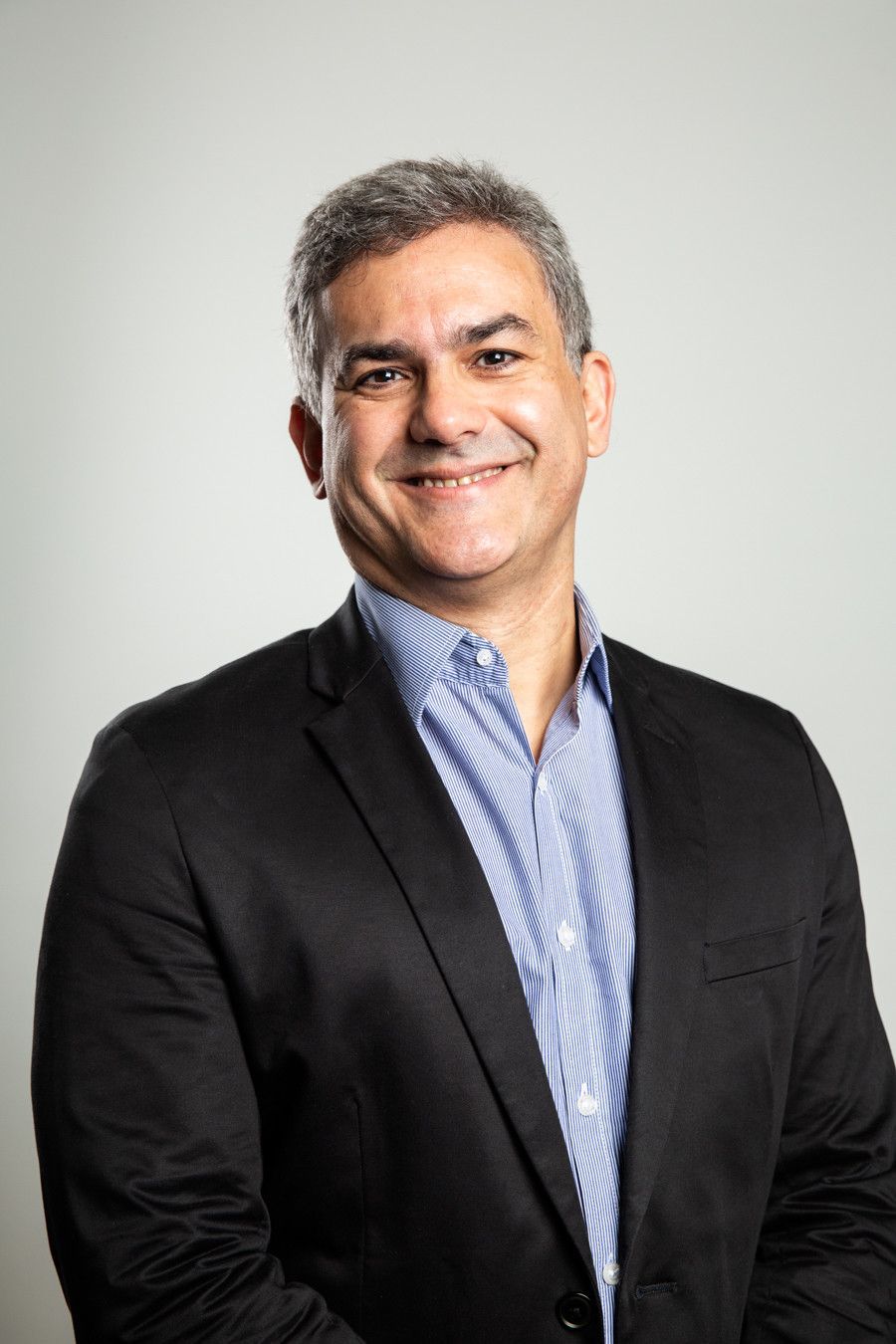 Homem sorridente de blazer preto e camisa azul contra um fundo cinza claro.