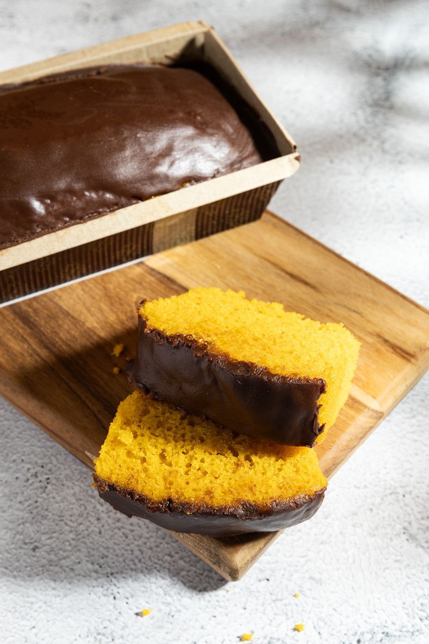 Duas fatias de bolo amarelo com cobertura de chocolate sobre uma tábua de madeira ao lado de um bolo em uma forma.