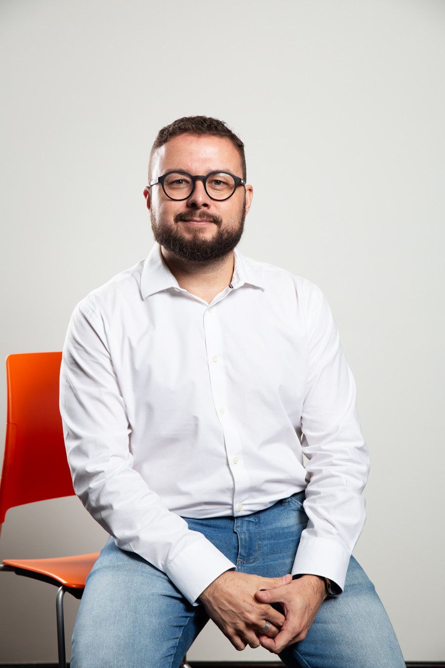 Homem sentado, vestindo camisa branca e calça jeans, ao lado de uma cadeira laranja contra uma parede clara.