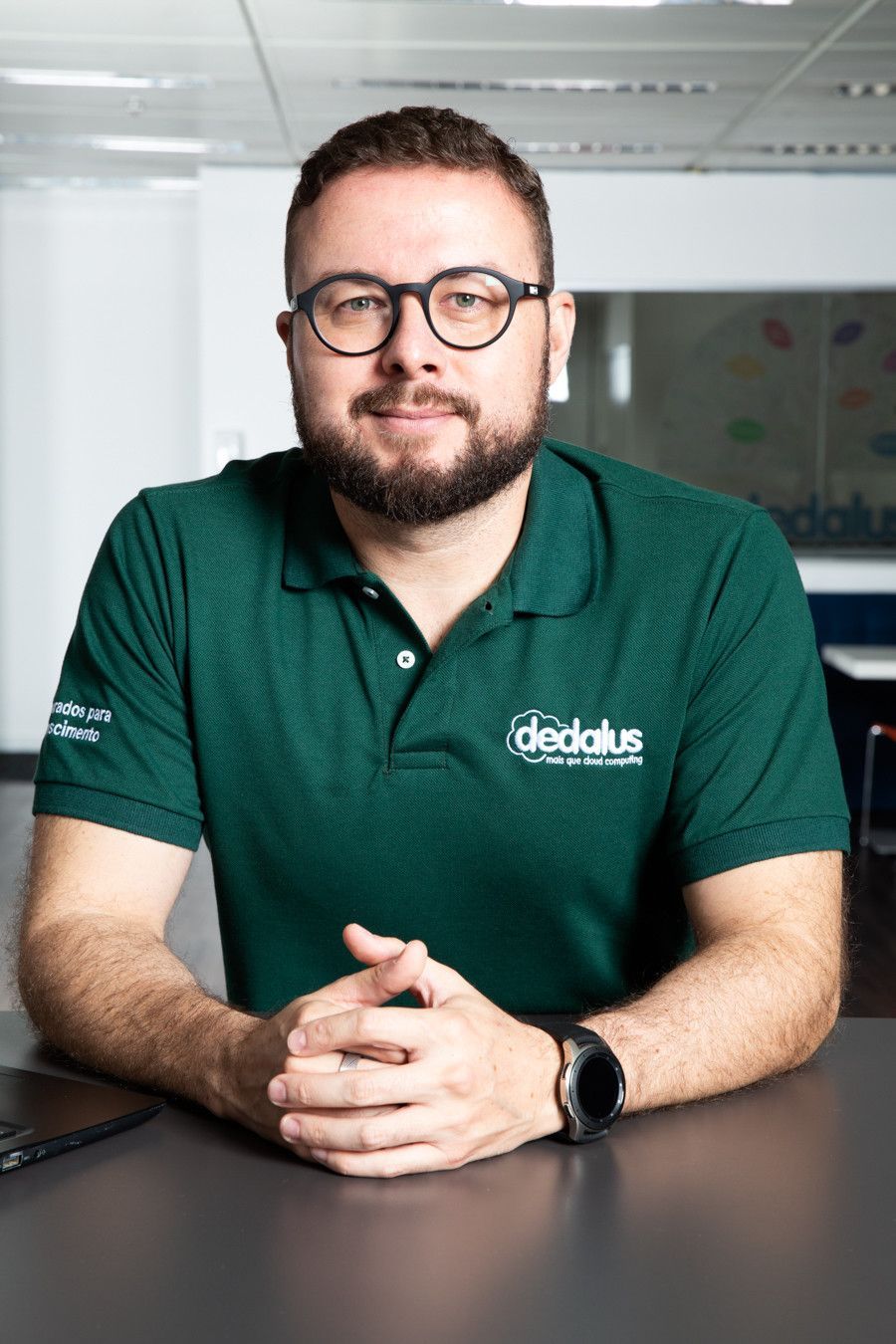 Pessoa vestindo uma camisa polo verde e óculos, sentada à mesa em um escritório.