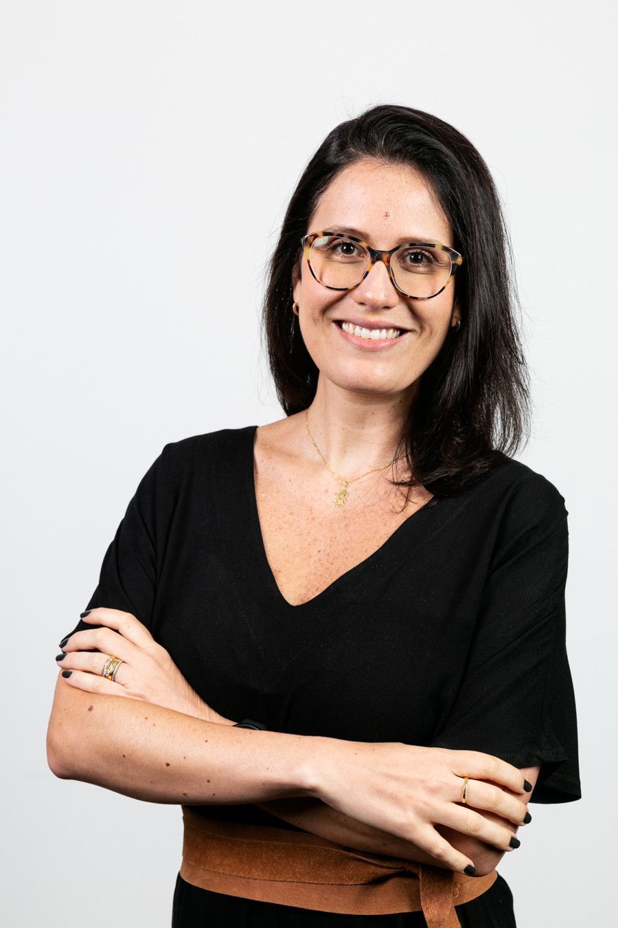 Pessoa sorridente de óculos, vestindo blusa preta, com os braços cruzados, contra um fundo branco.
