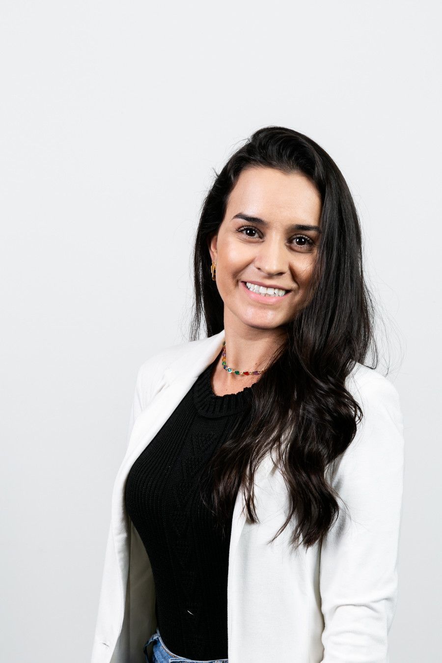 Mulher sorridente vestindo blazer branco e blusa preta, em pé contra um fundo cinza claro.