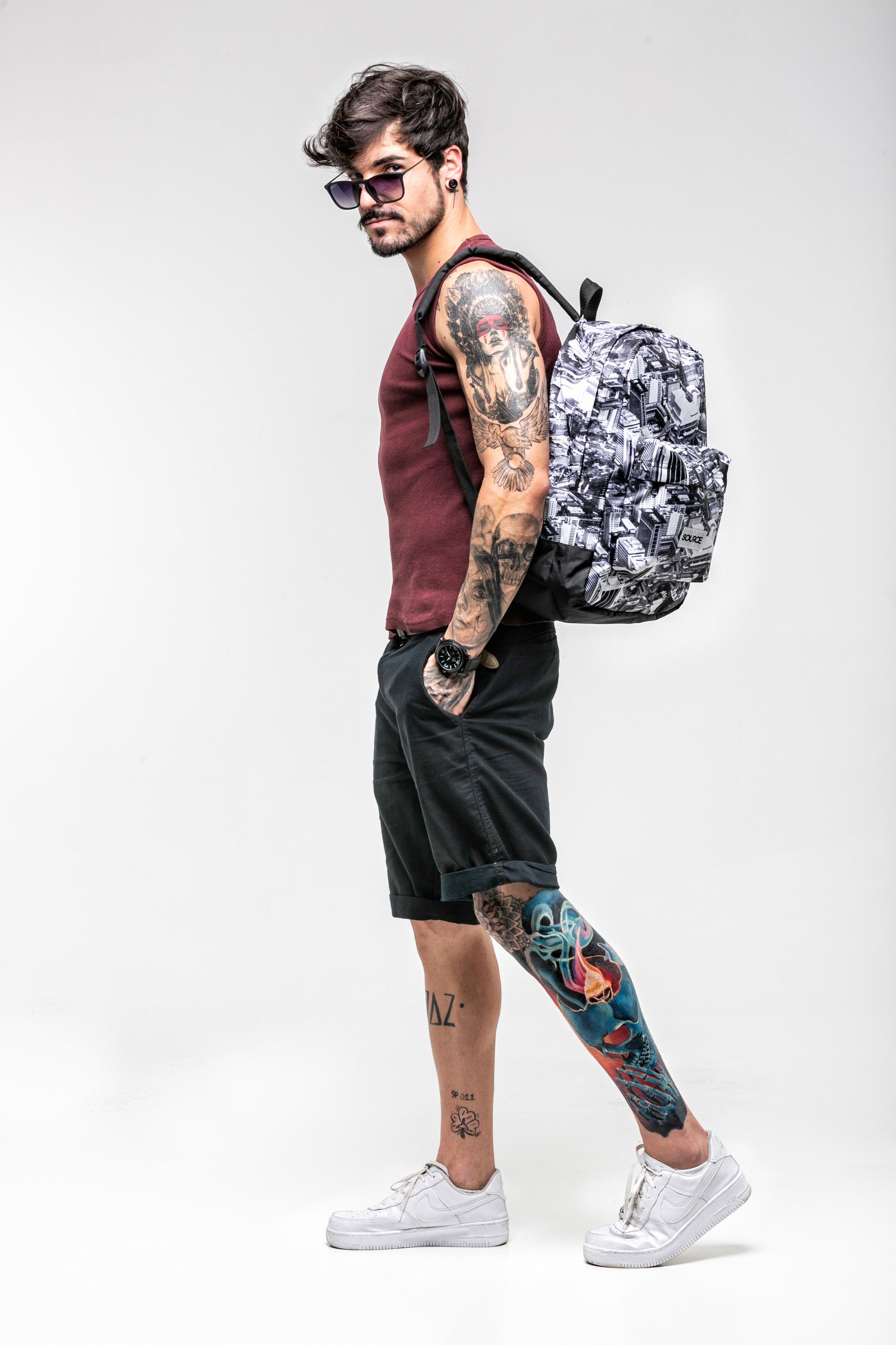 Homem tatuado vestindo regata bordô, bermuda preta e tênis brancos, carregando uma mochila estampada.