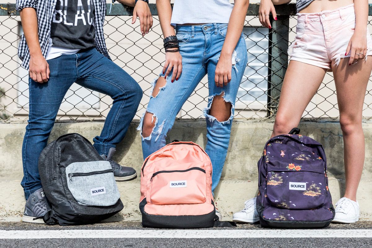 Três pessoas em pé atrás de três mochilas em uma calçada, com uma cerca de arame ao fundo.