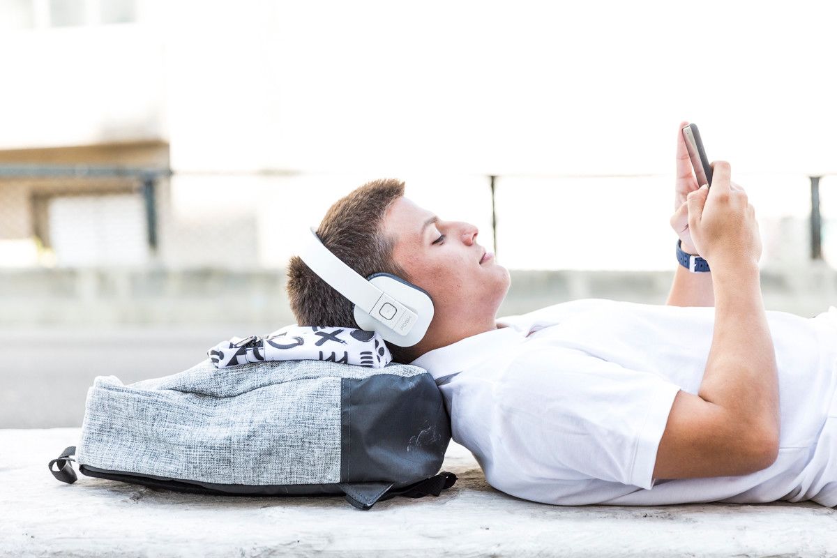 Pessoa deitada em um tapete no chão, usando fones de ouvido e um smartphone, com uma mochila ao lado.