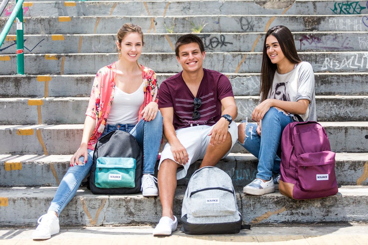Três estudantes sentam-se em degraus externos com mochilas e cadernos, sorrindo para a câmera.