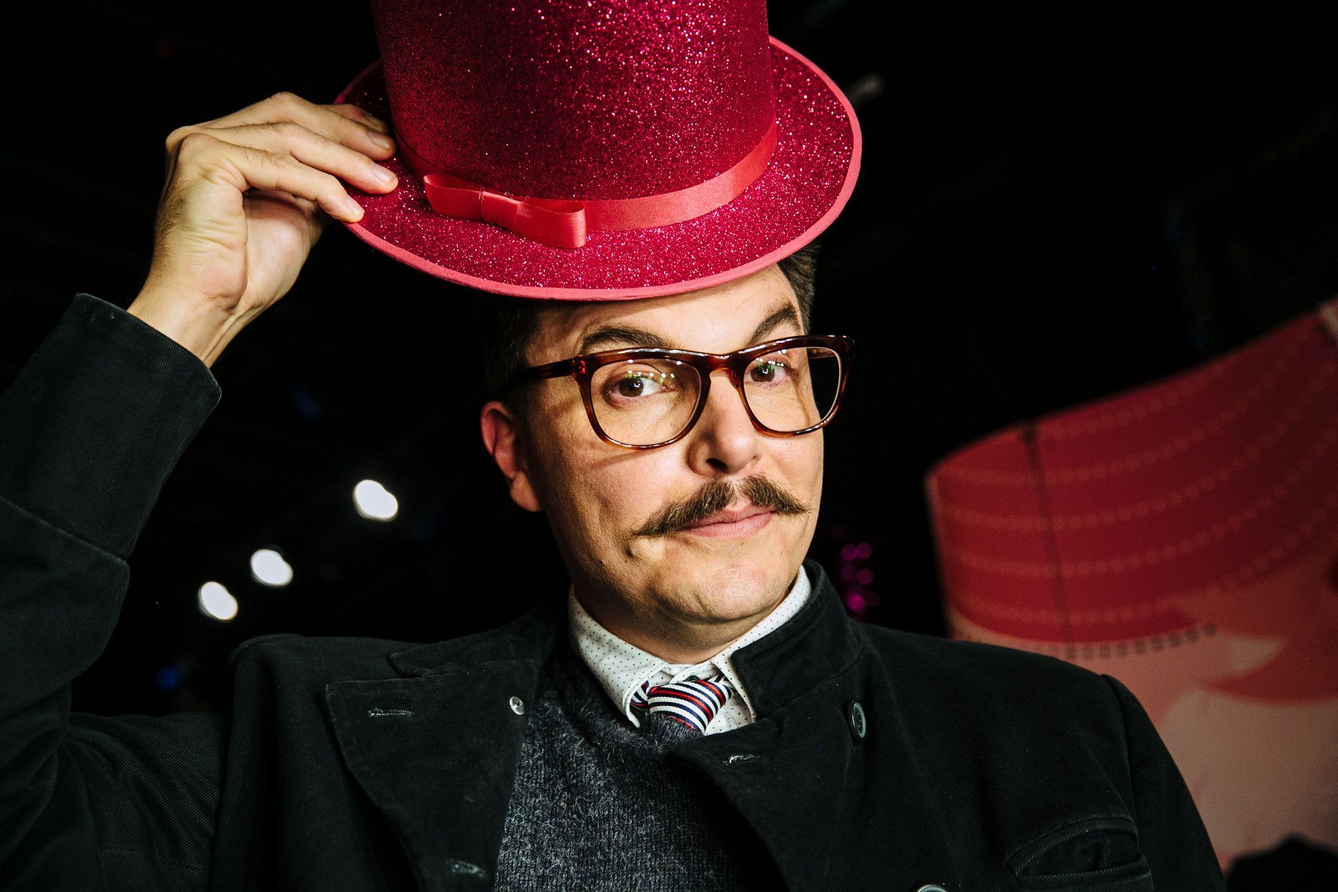 Pessoa de óculos inclinando uma cartola rosa brilhante com glitter contra um fundo escuro.