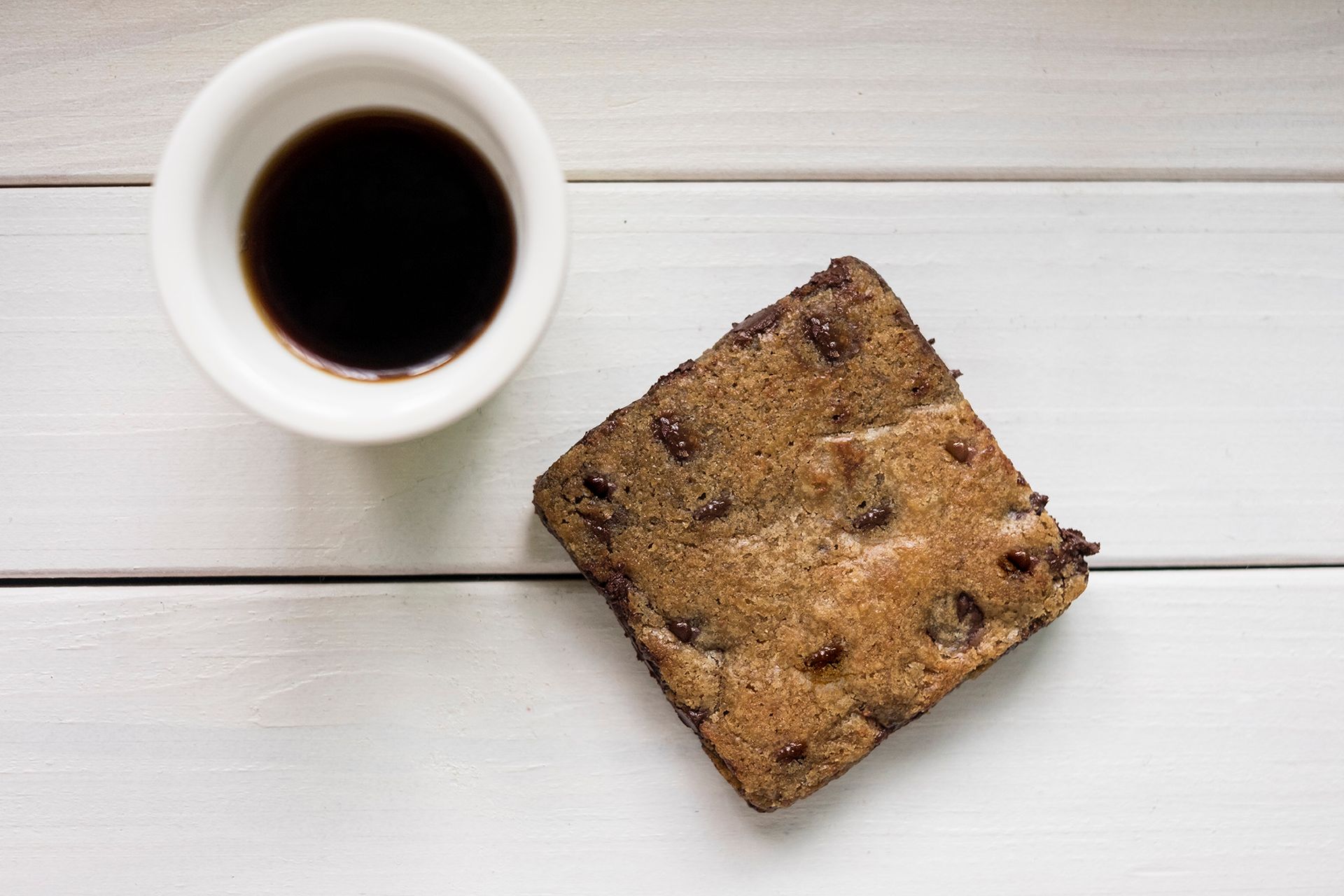 Xícara de café preto ao lado de um biscoito quadrado com gotas de chocolate em uma mesa de madeira branca.