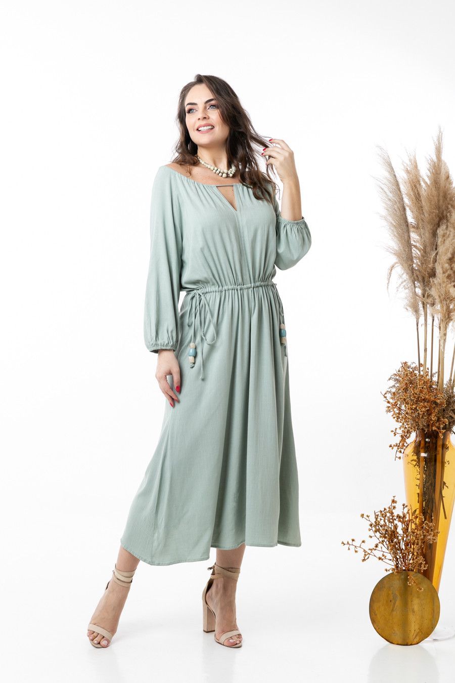 Modelo em um vestido midi verde-sálvia com cinto, posando ao lado de capim-dos-pampas seco sobre um fundo branco.