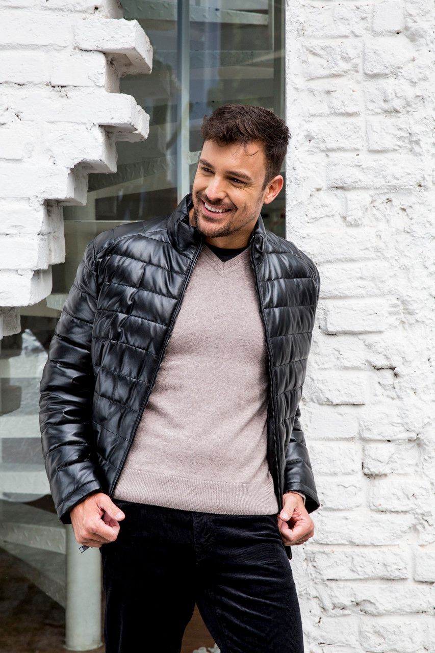 Homem de jaqueta preta e suéter bege parado junto a uma parede de tijolos brancos, sorrindo e olhando para o lado.