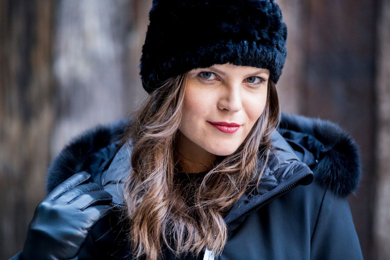 Mulher com chapéu e casaco de pele pretos, sorrindo ao ar livre em um cenário de inverno.