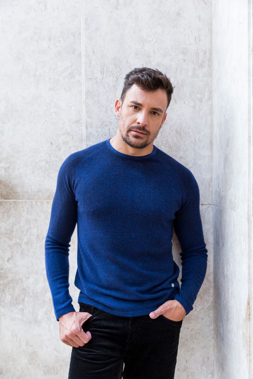Homem de suéter azul e calça preta, em pé contra uma parede clara, com as mãos nos bolsos.