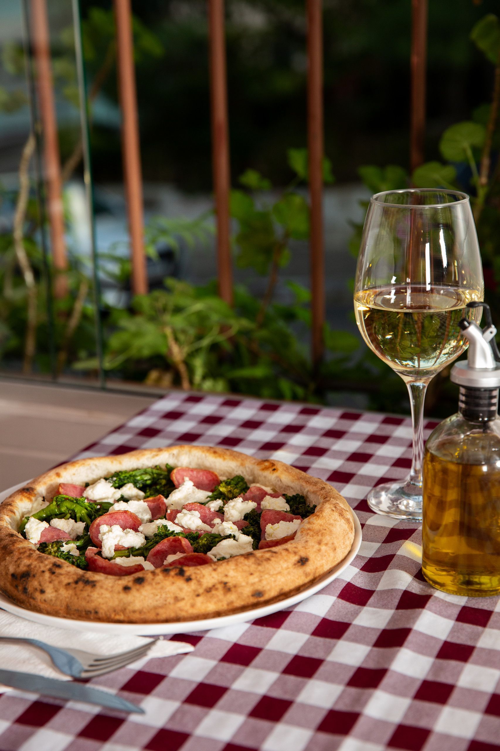 Pizza e um copo de vinho branco sobre uma toalha de mesa xadrez na área externa de um café.
