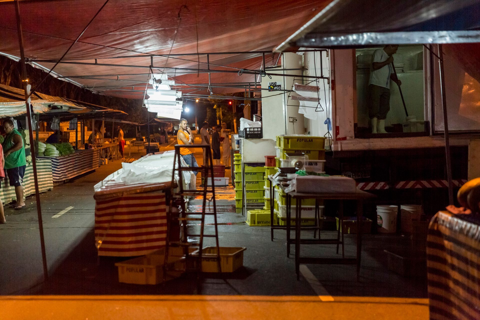 Corredor de mercado interno pouco iluminado, com pilhas de caixas, mesas e luzes quentes no teto vermelho.
