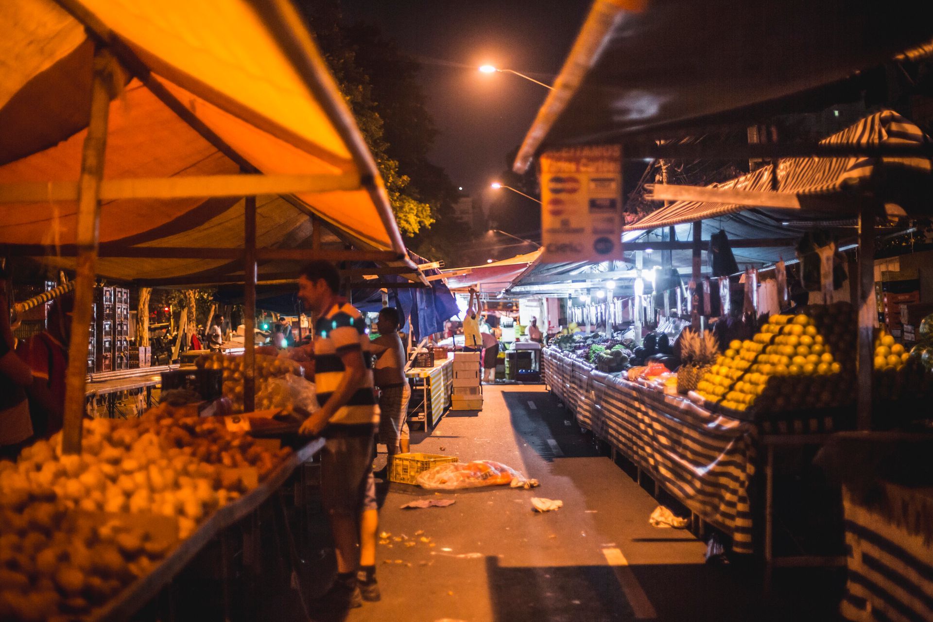 Mercado de rua noturno com barracas de produtos agrícolas sob toldos laranja e luzes brilhantes.