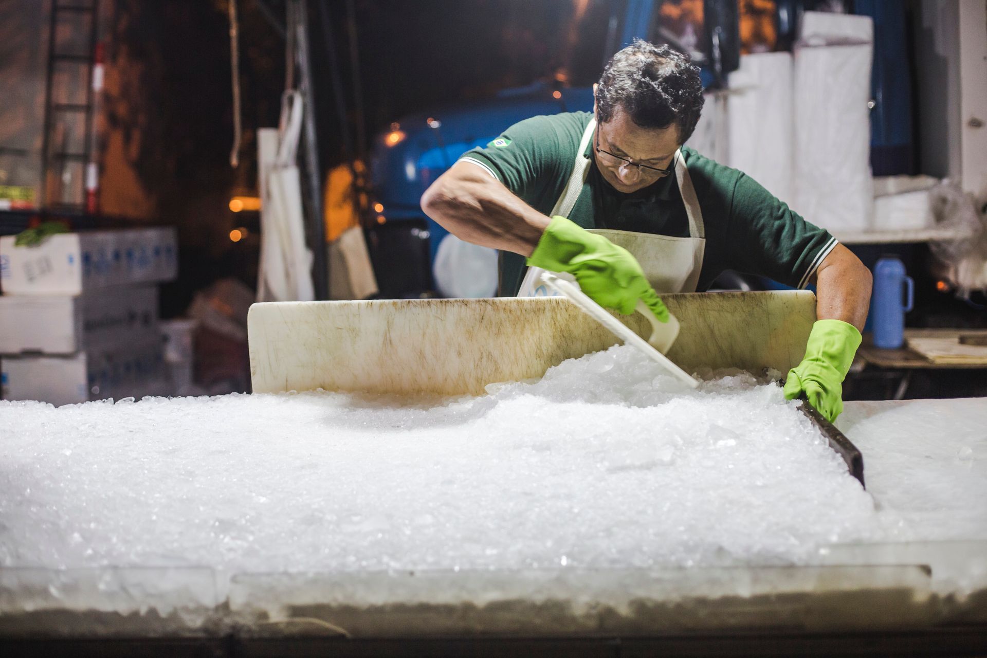 Trabalhador com luvas verdes a remover gelo de uma banca de mercado de peixe.