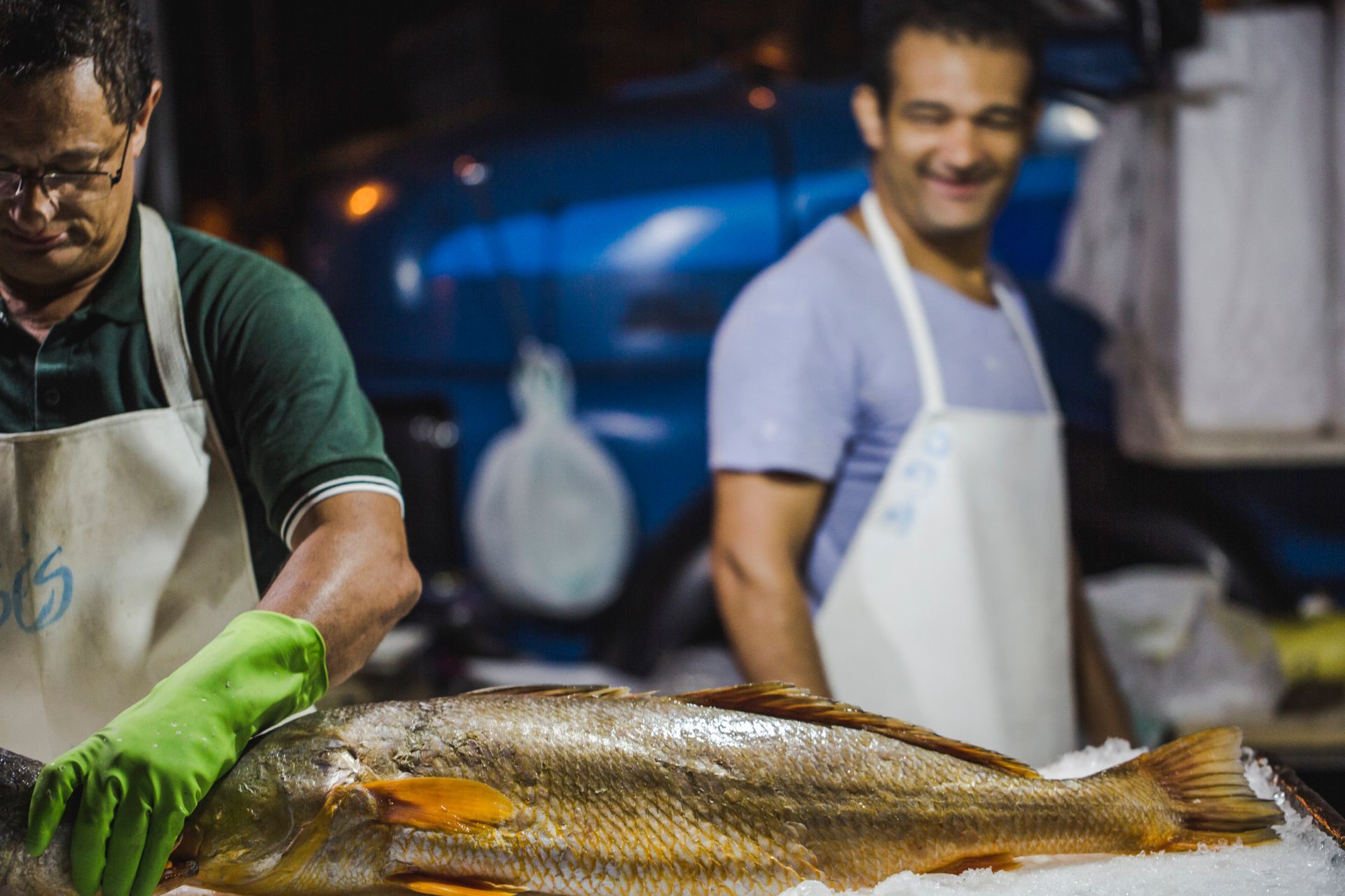 Peixeiro de luvas verdes preparando um peixe inteiro grande em uma barraca de mercado, com outro trabalhador ao fundo.