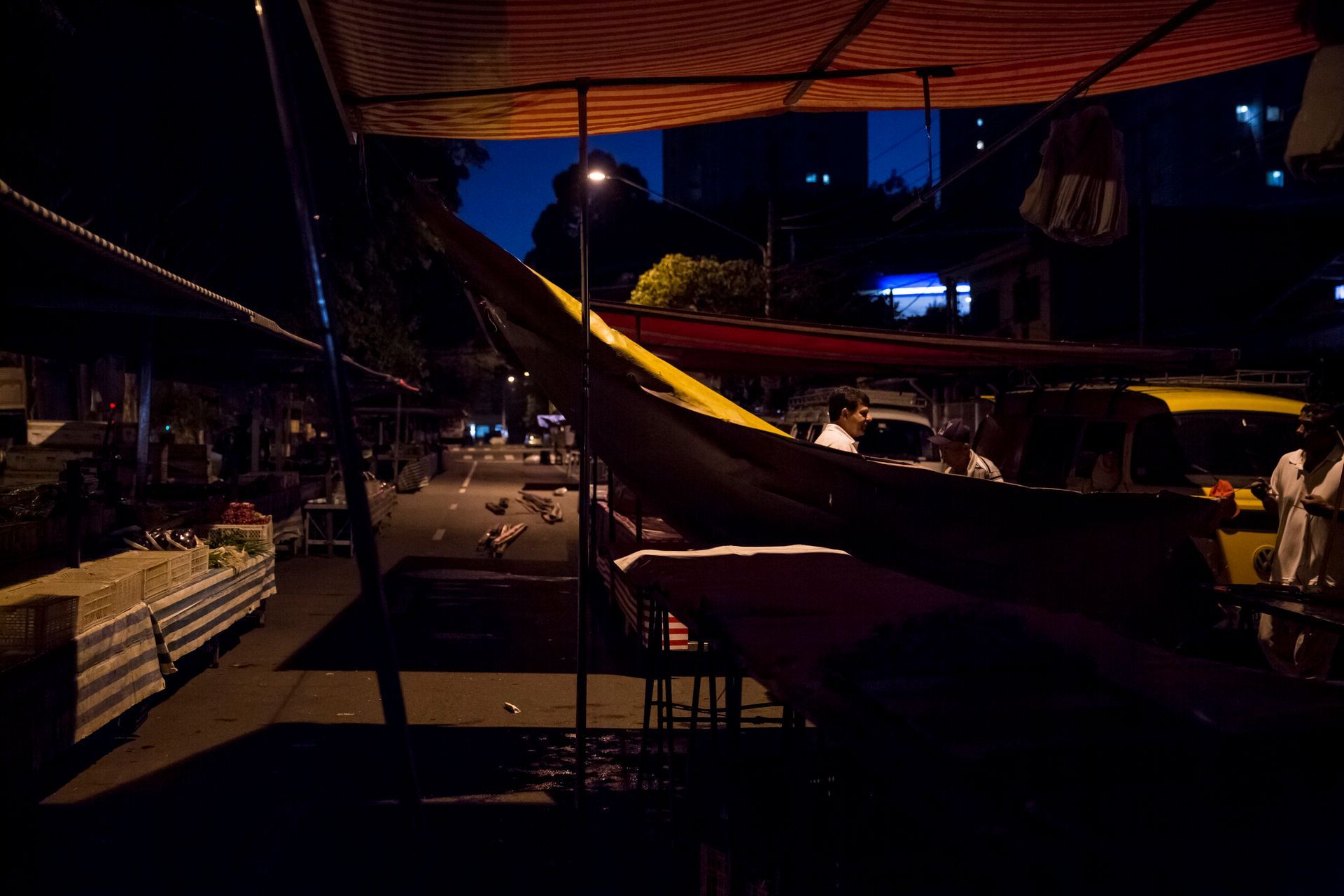 Mercado ao ar livre com mesas sob um toldo ao entardecer, iluminado por luzes quentes e pelo céu azul da noite.