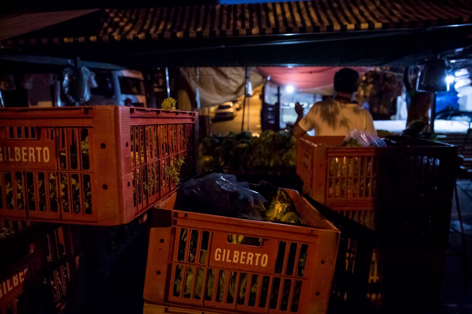 Barraca de mercado noturno com caixas de laranjas e pessoas sob um telhado de telhas pouco iluminado por luzes quentes.