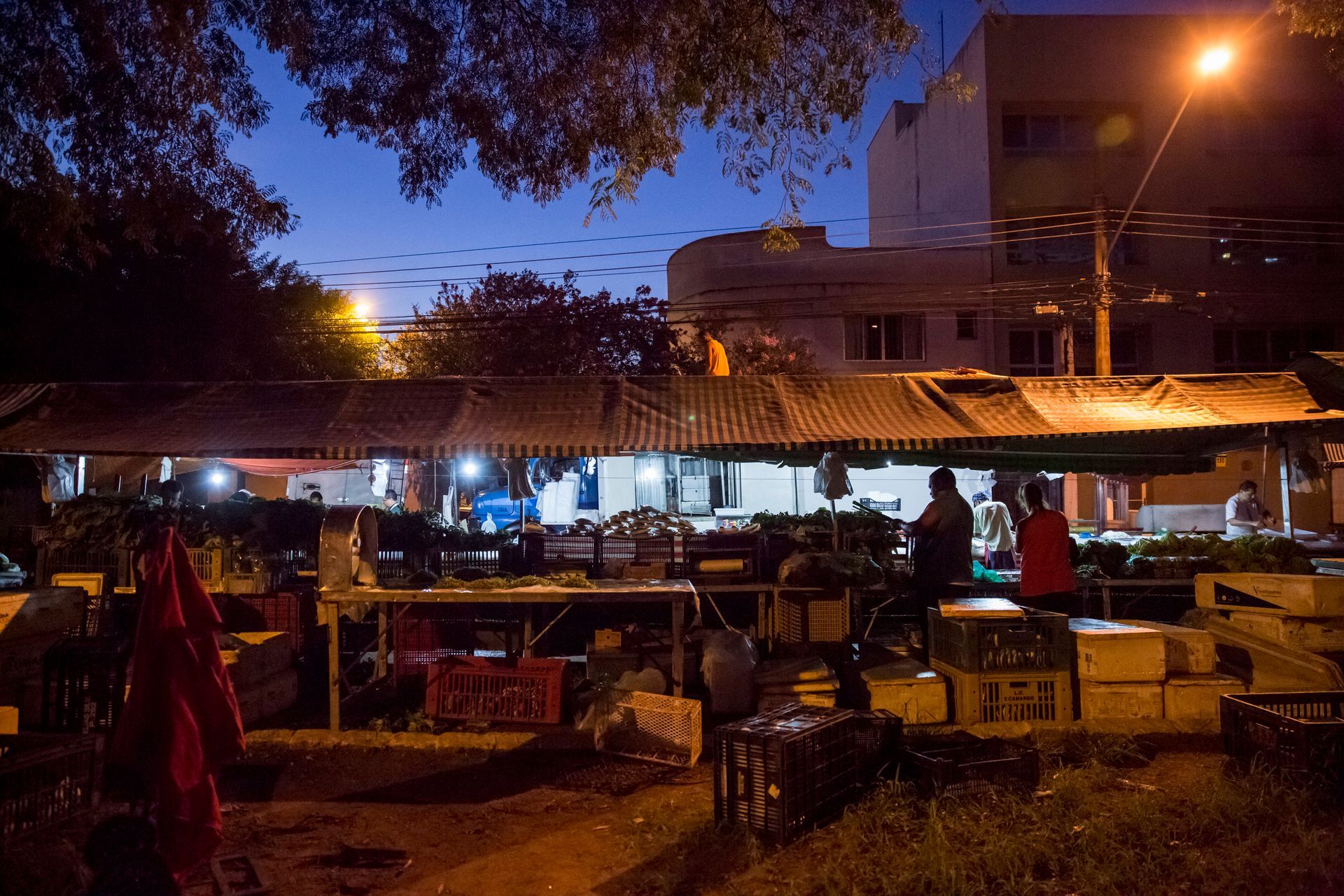 Mercado noturno ao ar livre com barracas iluminadas, mesas e pessoas sob um telhado de zinco, sob a luz quente dos postes.