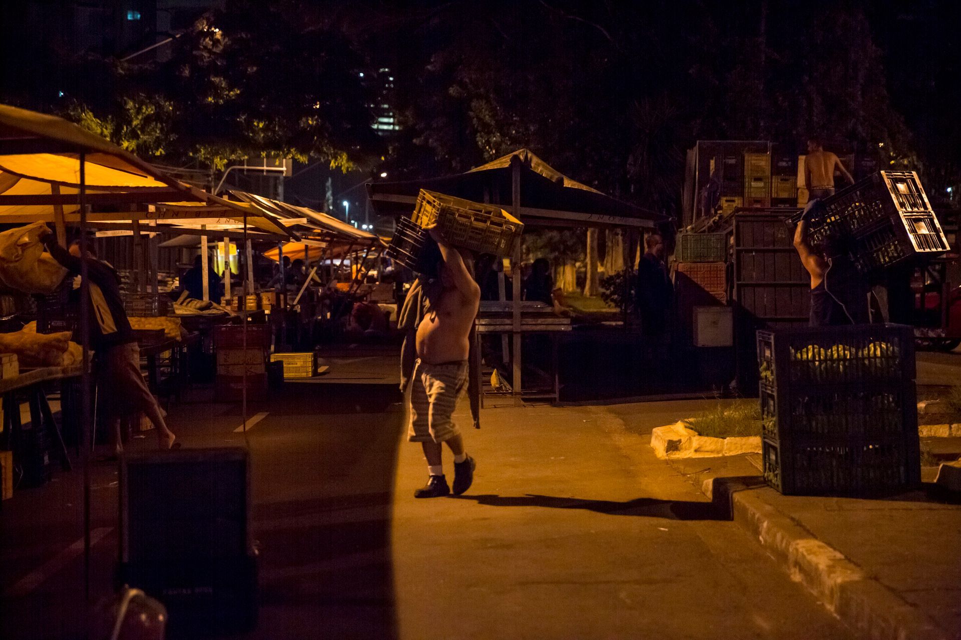 Calçadão de um mercado noturno iluminado por luzes amarelas e aconchegantes, com uma pessoa em pé entre barracas e guarda-chuvas.