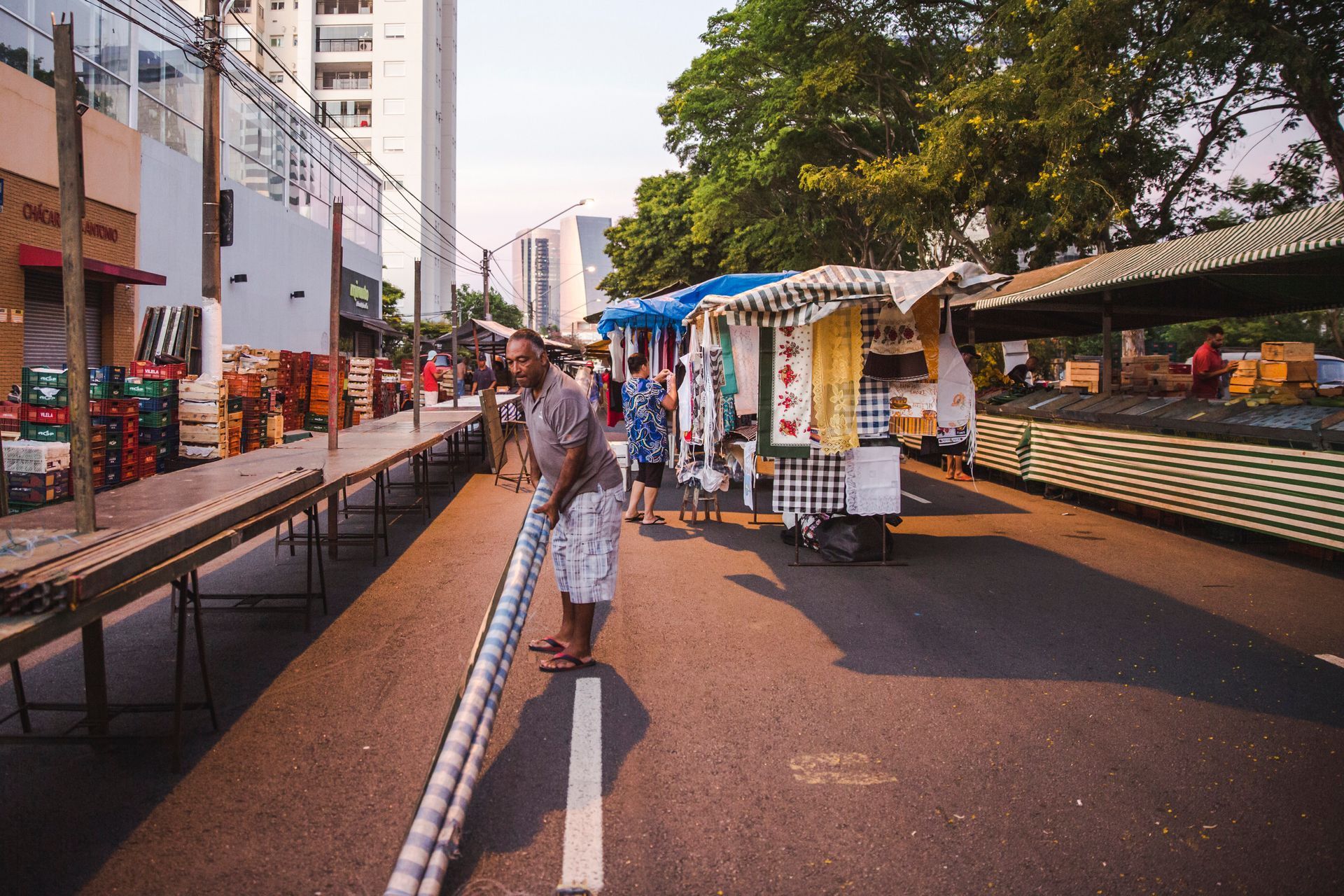 Rua de mercado para pedestres com barracas, uma pessoa caminhando no centro e árvores margeando a via.
