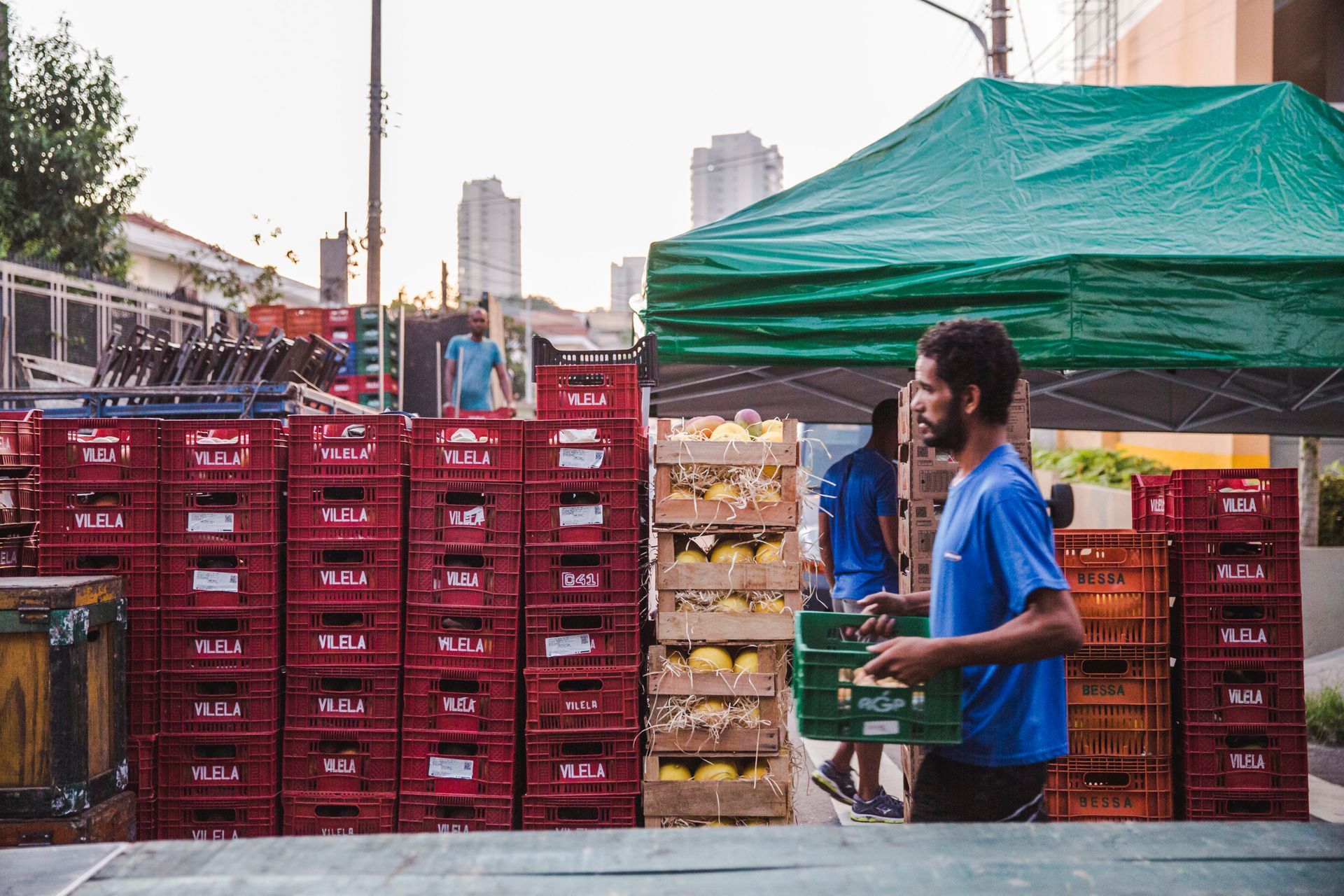 Pessoa carregando caixas em um mercado ao ar livre, ao lado de caixas vermelhas de produtos agrícolas empilhadas e uma tenda verde.