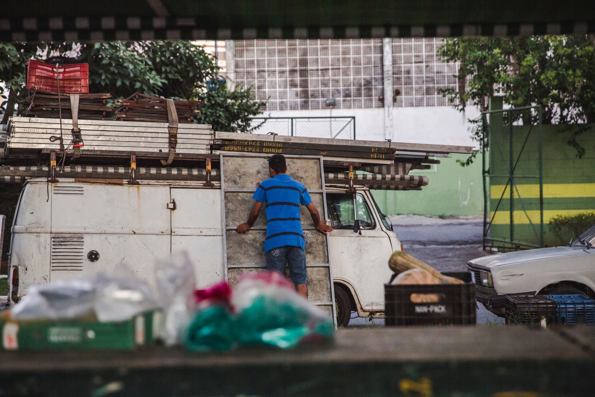 Vendedor ambulante em pé ao lado de um caminhão branco sob árvores, com sacolas e caixas em primeiro plano.