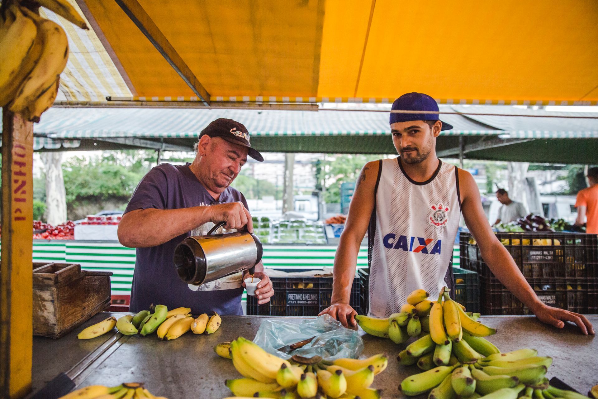 Dois homens em uma banca de frutas, um servindo suco enquanto bananas estão dispostas no balcão.
