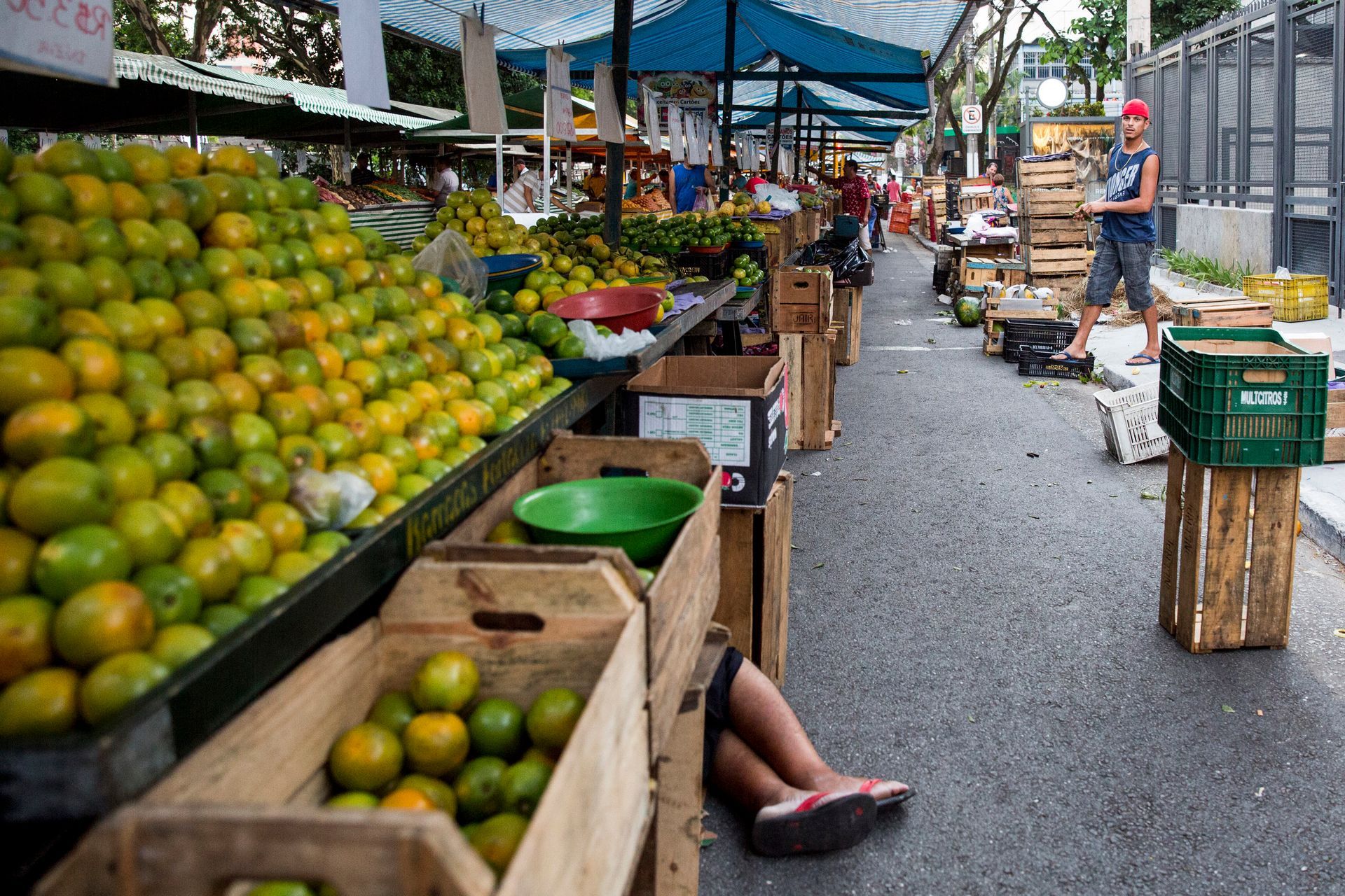 Mercado ao ar livre com frutas cítricas e outros produtos empilhados em barracas de madeira ao longo de uma passarela molhada.