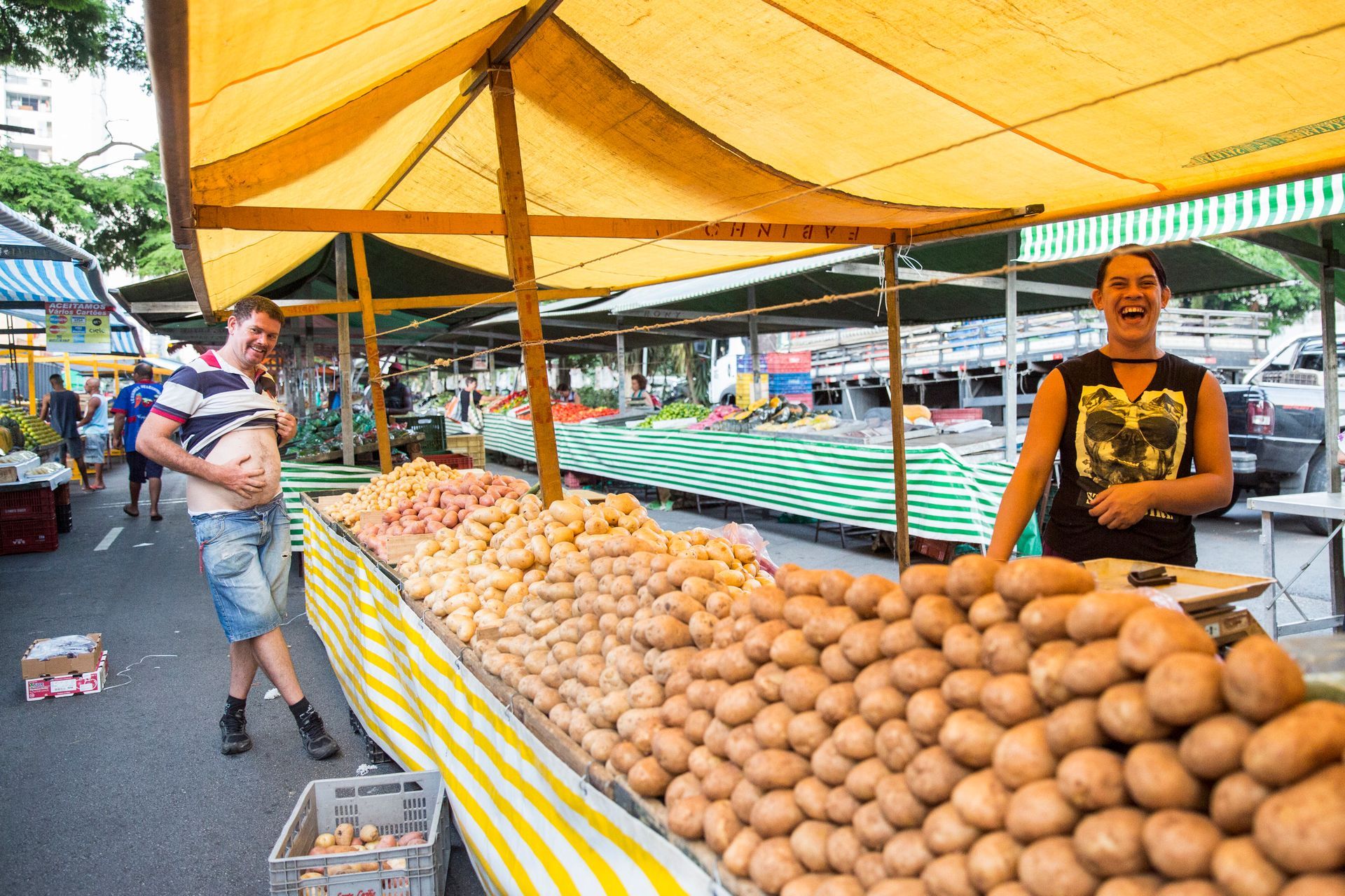 Banca de mercado com pilhas de cocos sob um toldo amarelo, vendedor sorrindo ao lado da exposição.