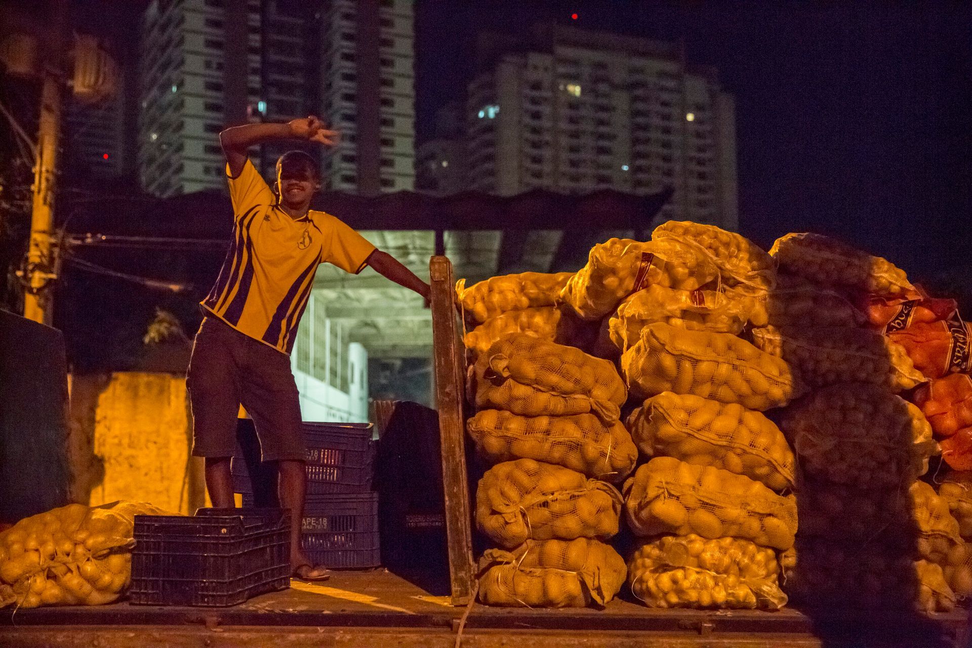 Trabalhador empilhando sacos em um carrinho à noite, sob a luz de postes, com prédios altos ao fundo.